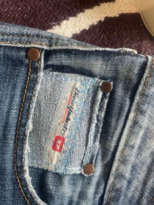 Blå slitna jeans från Diesel - Säljer ett par klassiska blå jeans från Diesel med snyggt sliten tvätt och kontrastsömmar. Jeansen har fem fickor, bälteshällor och knappgylf. Perfekta för en avslappnad och trendig look.