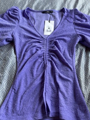 Lila topp med puffärm och rynk - Har andra storlekar,XS,S,M,L Trendig lila topp med v-ringning, puffärmar och snygg rynkdetalj framtill. Tillverkad i ett strukturerat syntetmaterial som ger en cool textur. Perfekt för dig som vill sticka ut med färg och form!