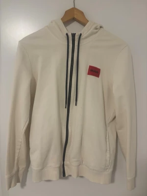 Vit hoodie med dragkedja från Hugo set - Vit hoodie från Hugo med svart dragkedja och snörning i huvan. Klassisk passform med ribbade muddar och en röd Hugo-logga på bröstet. Perfekt för en clean och stilren look.