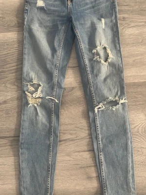 Blå slitna skinny jeans med hål - Säljer ett par blå skinny jeans med stora slitningar och hål på både lår och knän. Jeansen har en klassisk femficksmodell och dragkedja fram. Perfekta för en avslappnad och trendig streetstyle-look. 28 midjan 30 längd 