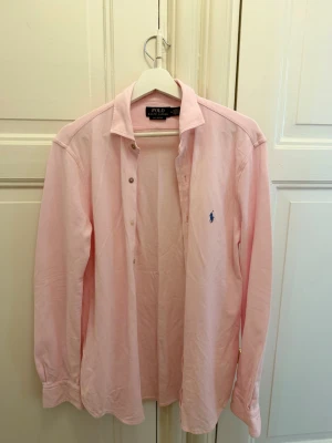 Ljusrosa skjorta från Polo Ralph Lauren - Säljer en ljusrosa skjorta från Polo Ralph Lauren i modellen Knit Oxford. Skjortan har klassisk krage, långa ärmar och knappar framtill. Ikoniska blå loggan broderad på bröstet. Perfekt för dig som gillar stilrena plagg med en preppy vibe.