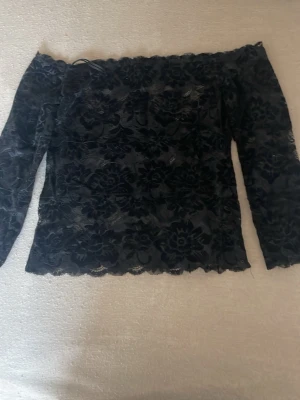 Svart spetsblus offshoulder Guess - Säljer en svart offshoulder blus från Guess i storlek M. Blusen är täckt av blommig spets och har långa ärmar. Perfekt för dig som vill ha en elegant och trendig topp med snygga detaljer.