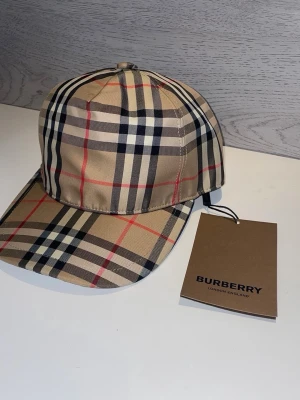 Beige keps från Burberry med rutmönster - Snygg beige keps från Burberry med det klassiska rutmönstret i svart, vitt och rött. Kepsen har böjd skärm och justerbar rem bak. Tillverkad i ett slätt tyg som ger en exklusiv känsla. Perfekt accessoar för att lyfta din outfit.