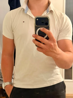 Vit pikétröja från Tommy Hilfiger - Klassisk vit pikétröja från Tommy Hilfiger med korta ärmar och blå detalj på kragen. Tröjan har en liten logga på bröstet och är tillverkad i mjuk bomull. Den är i storlek 164 vilket motsvarar ca XS, skriv gärna för frågor eller funderingar!