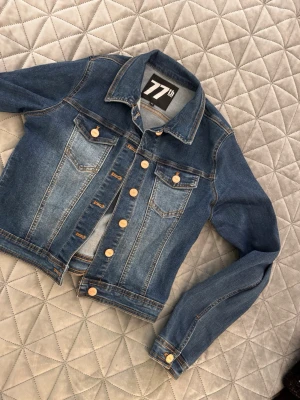 Blå jeansjacka från 77th - Snygg blå jeansjacka från 77th i klassisk modell med två bröstfickor och guldfärgade knappar. Jackan har en cool tvättad look med kontrastsömmar och är perfekt för dig som vill ha en tidlös och trendig stil.