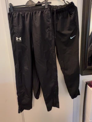 Svarta träningsbyxor Nike & Under Armour - Två par svarta träningsbyxor, ett från Nike och ett från Under Armour. Båda har resår i midjan och vid benslut, samt logga på vänster ben. Perfekta för gymmet eller löprundan. Materialet är lätt och andas bra.