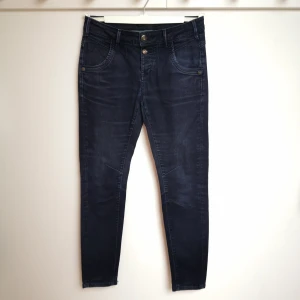Mörkblå jeans från Mos Mosh - Mörkblå jeans från Mos Mosh med dubbla knappar i midjan och klassiska fem fickor. Byxorna har en mjuk sammetsyta. Jeansen har en diskret tvättad look, detaljerade sömmar och ett litet guldfärgat emblem på bakfickan. Perfekt passform och stretchigt material för en cool stil. Storlek W27 en längd. 