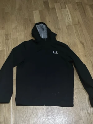 Svart vindjacka med huva Under Armour - Snygg svart vindjacka från Under Armour med huva och dragkedja. Jackan har lång ärm och en minimalistisk logga på bröstet. Perfekt för blåsiga dagar och enkel att matcha med olika outfits.