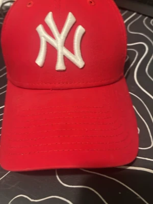Röd New Era NY keps - Snygg röd keps från New Era med klassisk böjd skärm och broderad vit NY-logga framtill. Kepsen har ventilerande hål och justerbar passform. Materialet är troligtvis bomull eller bomullsblandning. Perfekt för dig som gillar streetwear och sportig stil.