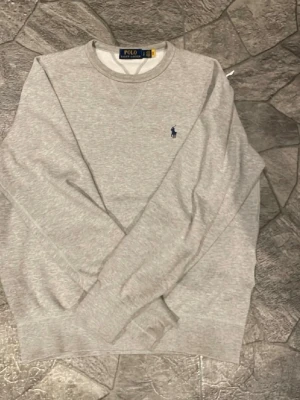  Polo Ralph Lauren tröja | S - Klassisk grå sweatshirt från Polo Ralph Lauren med rund halsringning och den ikoniska mörkblå broderade loggan på bröstet. Tröjan har ribbade muddar vid ärmslut och nederkant. Perfekt för en avslappnad och stilren look.