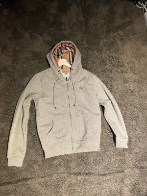 Grå hoodie med dragkedja och huva - Snygg grå hoodie  med dragkedja framtill och klassiskt rutigt foder i huvan. Hoodien har dragsko i huvan, två fickor fram och diskret logga broderad på bröstet. Tillverkad i mjukt bomullsmaterial för en bekväm känsla.