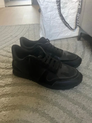 Svarta sneakers valentino rockrunners - Säljer ett par helsvarta sneakers med snygga mockadetaljer och klassisk snörning. Skorna har en låg profil och en stilren design som passar till det mesta. Perfekta för dig som gillar en clean och enkel look.