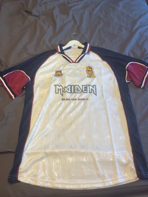 Vit och vinröd Iron Maiden fotbollströja ”Limited” - Säljer en unik vit fotbollströja med vinröda och marinblå detaljer. Tröjan har Maiden- och Brave New World-tryck på bröstet, samt nummer 7 på ryggen. Kragen har Maiden-tryck och materialet är glansigt polyester. Perfekt för dig som gillar sport och musik.