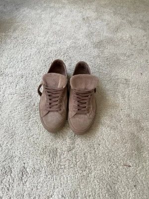 Bruna låga sneakers i mocka - Säljer ett par stilrena bruna sneakers i mocka med perforerad plös och matchande bruna skosnören. Skorna har en klassisk låg siluett och en ton-i-ton yttersula. Perfekta för dig som gillar en clean och enkel look.Frakt bitalas av köparen