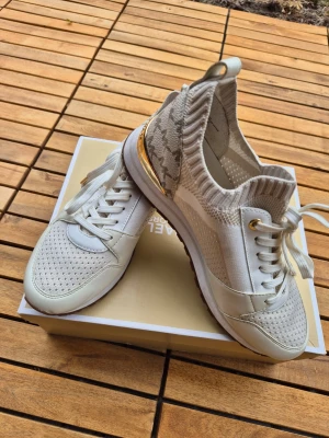 Beige sneakers med gulddetaljer - Snygga beige sneakers med stickad ovandel, meshpaneler och guldfärgade detaljer vid hälen. Skorna har vita snören, vit sula och brunt mönstrad yttersula. Perfekt för dig som gillar en sportig men trendig look. Endast använda en gång 