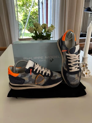 Philippe Model sneakers - Snygga sneakers från Philippe Model med camouflagemönster i blått och grått, detaljer i orange och vitt samt vita skosnören. Skorna har lågt skaft och en mix av mocka och textilmaterial. Perfekta för dig som gillar streetstyle och unika detaljer. Storlek 41, använd fåtal gånger! Allt på bilden tillkommer, låda och dustbag.