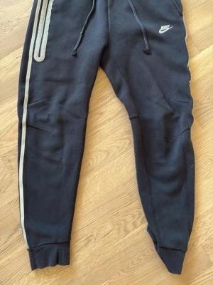 Svarta mjukisbyxor från Nike - Snygga svarta mjukisbyxor från Nike med vita detaljer och logga på vänster ben. Byxorna har dragsko i midjan, muddar vid bensluten och en vit rand längs sidan. Perfekta för chill eller träning, tillverkade i mjukt bomullsmaterial.