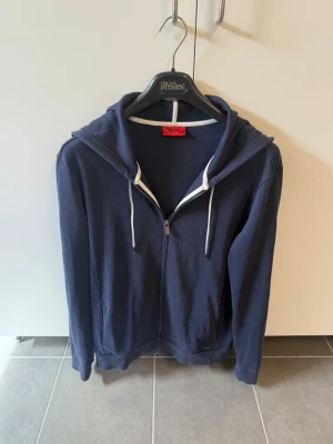 Mörkblå hoodie med dragkedja från Hugo Boss, stl M - Snygg mörkblå hoodie med dragkedja framtill och vita dragsnören i huvan. Klassisk modell med fickor på sidorna och ribbade muddar vid ärmslut och nederkant. Perfekt för en avslappnad och stilren look.
