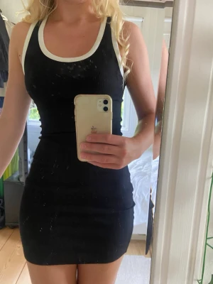Svart ärmlös ribbad klänning - Snygg svart kortklänning med vita ribbade kanter vid hals och ärmhål. Klänningen är ärmlös och har en kroppsnära passform som ger en sportig och stilren look. Perfekt för varma dagar eller att styla med en jacka.