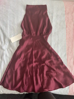 Vinröd halterneckklänning i satin - Snygg vinröd klänning i glansigt satinmaterial med halterneck och utsvängd kjol. Perfekt för dig som vill ha en elegant look med en modern twist. Klänningen har en smickrande passform och är ärmlös.