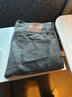 Grå Levi's 501 raka jeans - Säljer ett par klassiska grå Levi's 501 jeans med rak passform och femficksdesign. Jeansen har den ikoniska läderlappen bak i midjan och är tillverkade i slitstark bomull. Perfekta för dig som gillar tidlös stil och vill ha ett par jeans som funkar till allt.