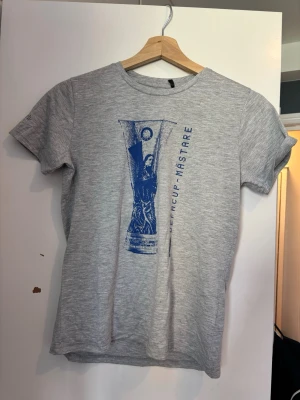 Grå ifk Göteborg T-shirt med blått tryck  - Snygg grå t-shirt med ett stort blått tryck av en pokal och texten 'CUP-MÄSTARE' på framsidan. T-shirten har rund hals och korta ärmar. Tillverkad i mjukt material som känns skönt mot huden. Perfekt för dig som gillar sportig och avslappnad stil. Nästan aldrig använd