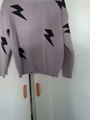 Ljuslila stickad tröja med blixtar - Cool ljuslila stickad tröja med svarta blixtmotiv. Tröjan har rund halsringning, långa ärmar och ribbstickade muddar vid ärmslut och nederkant. Perfekt för dig som gillar unika detaljer och vill sticka ut.