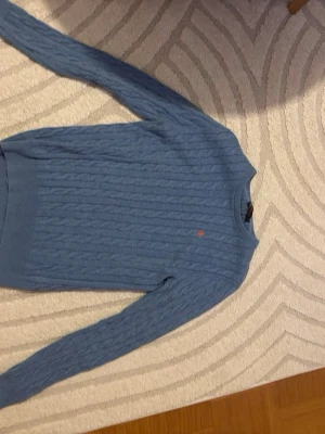 Blå kabelstickad tröja från Morris - Snygg blå kabelstickad tröja från Morris med rund hals och diskret logga broderad på bröstet. Tröjan har långa ärmar och ribbade muddar vid ärmslut och nederkant. Perfekt för dig som gillar klassisk stil. En fin och rik känsla då hoodien är lite Stockholm stiligt