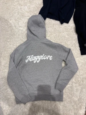 Grå hoodie från Maggiore - Snygg grå hoodie från Maggiore med klassisk huva och vita dragsnören. Hoodien har en stor magficka framtill och är tillverkad i mjukt material med borstad insida för extra komfort. Perfekt för en avslappnad och stilren look.