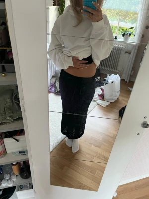 Svart stickad långkjol med snörning - Snygg svart långkjol i stickat material med dekorativa hål och snörning i midjan. Kjolen har en rak passform och är perfekt för dig som gillar en stilren och trendig look. Passar till många olika outfits och är superenkel att styla.