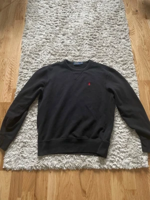 Svart sweatshirt från Polo Ralph Lauren - Klassisk svart sweatshirt från Polo Ralph Lauren med rund halsringning och ribbade muddar. Tröjan har en liten röd broderad logga på bröstet och är tillverkad i mjukt bomullsmaterial. Perfekt för en avslappnad och stilren look.
