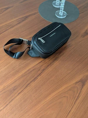 Svart axelväska från Valentino - Snygg svart axelväska från Valentino med justerbar axelrem och silverfärgade dragkedjor. Väskan har en rektangulär form, flera fack och diskret logga på framsidan. Materialet är slätt och remmen har präglad logotyp. Perfekt för att bära det viktigaste.