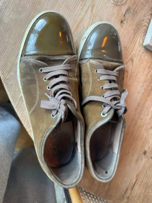 Bruna/gröna/oliv färgade lavin skor - Säljer ett par bruna sneakers med mockaliknande material och glansig, lackad tå i mörkbrunt. Skorna har beige sula och matchande bruna skosnören. Klassisk låg modell med rundad tå och metallfärgade öljetter för snörningen. De har ett litet ”glap” som visas i bilderna men inget som stör/ är ett stort problem alls. Samt ett pyttelitet hål i tyget vid hälen/ACL. Perfekt nu till hösten och skolstarten då det är riktigt fin färg. Kom så diskuterar vi pris