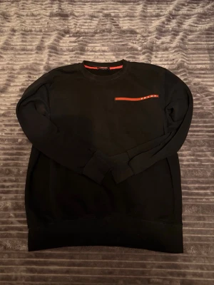 Svart sweatshirt från Prada - Säljer en svart sweatshirt från Prada med rund halsringning och en röd detalj med logga på bröstet. Tröjan har långa ärmar och ribbade muddar vid ärmslut och nederkant. Perfekt för dig som gillar stilrena designer och exklusiva märken.