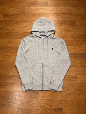Grå zip hoodie från Polo Ralph Lauren - Klassisk grå hoodie med dragkedja från Polo Ralph Lauren. Tröjan har huva med dragsko, två fickor framtill och den ikoniska lilla broderade loggan på bröstet. Tillverkad i mjukt sweatshirtmaterial som är perfekt för chill dagar. Tröjan är liten i storlek så den passar mer xs än s