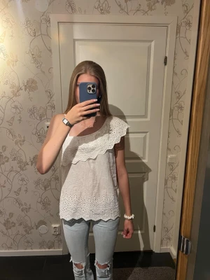Vit oneshoulder topp med volang - Säljer en söt vit oneshoulder topp med bred volang och broderade spetsdetaljer. Toppen är ärmlös och har ett luftigt, somrigt material som passar perfekt till jeans eller kjol. Perfekt för dig som gillar romantiska detaljer och unik design. Den har en ärm på andra sidan som man kan ha om man vill, man ser lite av den på första bilden men tröjan funkar lika bra utan bandet. Helt ny med lappen kvar. Det är storlek xxs men passar också xs/s för skulle säga att det är mer som en xs.