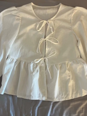 Vit blus med knytband och volang - Söt vit blus med tre knytband framtill och volang nedtill. Blusen har rund halsringning och långa puffärmar som ger en romantisk känsla. Tillverkad i ett lätt, vävt tyg som känns luftigt och skönt.