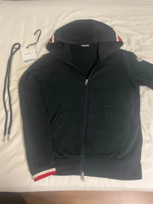 Svart Moncler hoodie med dragkedja - Snygg svart hoodie från Moncler med dragkedja framtill och huva. Detaljer i rött och vitt på huvan och ärmsluten ger en sportig vibe. Klassisk Moncler-logga på ärmen och känguruficka framtill. Perfekt för dig som vill ha en stilren och exklusiv hoodie.