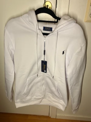 Vit hoodie från Polo Ralph Lauren - Snygg vit hoodie från Polo Ralph Lauren med dragkedja, huva och fickor framtill. Klassisk logga broderad på bröstet i svart. Tillverkad i mjukt bomullsmaterial och har justerbara snören i huvan. Perfekt för en clean och stilren look.