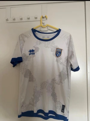 Vit Kosovo fotbollströja Erreà - Säljer en vit fotbollströja från Erreà med Kosovos landslagsemblem på bröstet. Tröjan har blå detaljer vid krage, ärmslut och nederkant samt ett grått mönster över hela plagget. Perfekt för dig som vill stötta Kosovo på match eller träning!