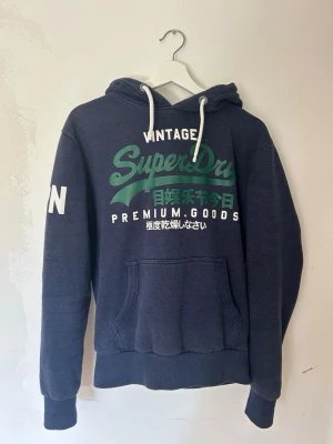 Mörkblå Superdry hoodie med tryck - Mörkblå hoodie från Superdry med stort grönt och vitt tryck på bröstet och japanska tecken. Klassisk känguruficka framtill, vita dragsnören i huvan och ribbade muddar. Perfekt för en avslappnad streetwear-look. Skriv till om ni är intresserade 😁