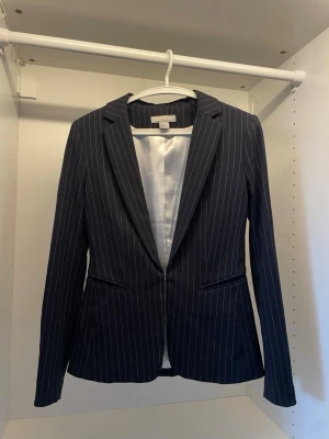Mörkblå kavaj med vita ränder - Snygg mörkblå kavaj med vita tunna vertikala ränder och klassisk krage. Kavajen har två fickor framtill och vit innerfoder. Perfekt för dig som vill ha en stilren och modern look. Passar till många olika outfits.