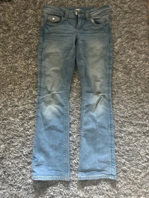 Ljusblå bootcut jeans med slitningar - Säljer ett par ljusblå bootcut jeans med slitningar vid knäna och klassiska fem fickor. Jeansen har en knappdetalj på fickan och är tillverkade i denim med en snygg tvätt. Perfekta för en avslappnad och trendig look. Storlek 158 från gina tricot.