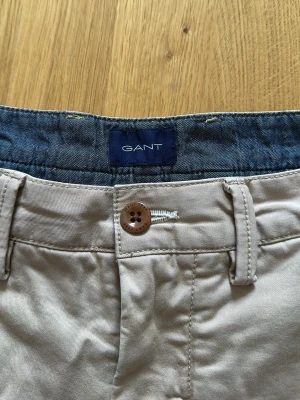 Beige shorts från GANT - Snygga beige shorts från GANT med klassisk design och uppvikta benslut. Tillverkade i bomull med knappstängning och bälteshällor. Perfekta för varma dagar och enkel att matcha med olika stilar. Lite solblekta