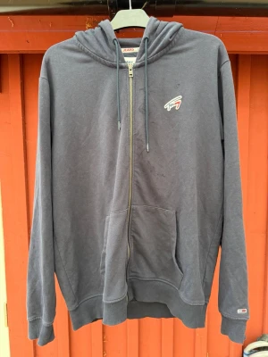 Mörkgrå hoodie från Tommy Jeans - Säljer en mörkgrå hoodie från Tommy Jeans med dragkedja, huva med dragsko och två stora fickor framtill. Liten broderad Tommy Jeans-logga på bröstet och en diskret logga på ärmen. Perfekt för en avslappnad och stilren look.