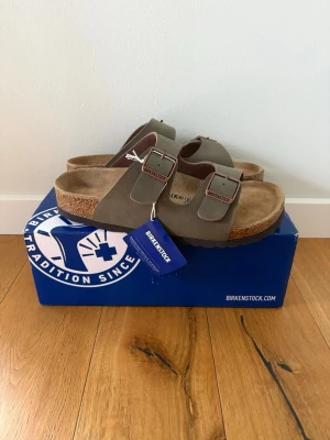 Birkenstock Arizona sandaler olivgrön - Säljer ett par Birkenstock Arizona sandaler i olivgrönt skinn med två breda remmar och justerbara metallspännen. Fotbädden är i mocka och sulan är i kork för extra komfort. Klassisk och tidlös modell som passar till det mesta.