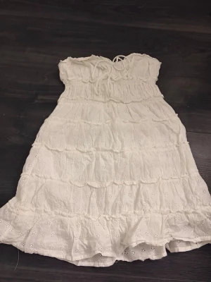 Vit kort klänning med spets och snörning - Söt vit kort klänning i bomull med broderade spetsdetaljer och volanger. Klänningen är axelbandslös med snörning upptill och har en luftig, romantisk känsla. Perfekt för varma dagar och sommarens alla tillfällen.
