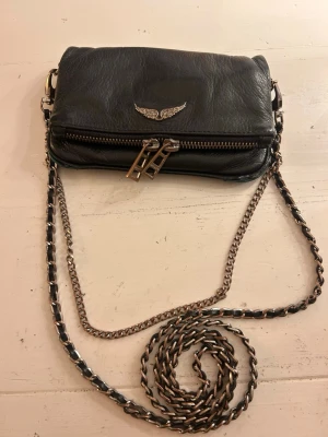 Äkta Zadig&voltaire väska  - Säljer nu min Zadig&voltaire väska då den inte kommer till användning. Den är väl använd (sliten) som man kan se på bild 4 och 5. Själva draget till kedjan på baksidan är av men man kan fortfarande stänga och öppna den (bild 6). Alla kedjor och själva märket har tappat sin färg lite också. Kom gärna privat om ni har frågor☺️Pris kan diskuteras💕