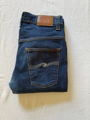 Mörkblå Nudie Jeans - Snygga mörkblå jeans från Nudie Jeans med klassisk femficksmodell och raka ben. Jeansen har kontrastsömmar och ikonisk brodyr på bakfickorna. Tillverkade i mjukt denimtyg som ger en skön passform och tidlös look. Storlek W31 L34!