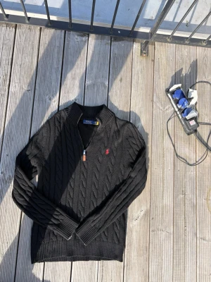 Svart kabelstickad tröja Polo Ralph Lauren - Kabelstickad svart tröja från Polo Ralph Lauren med röd broderad logga på bröstet. Tröjan har v-ringning med half zip och långa ärmar. Perfekt för dig som gillar klassisk och stilren design.
